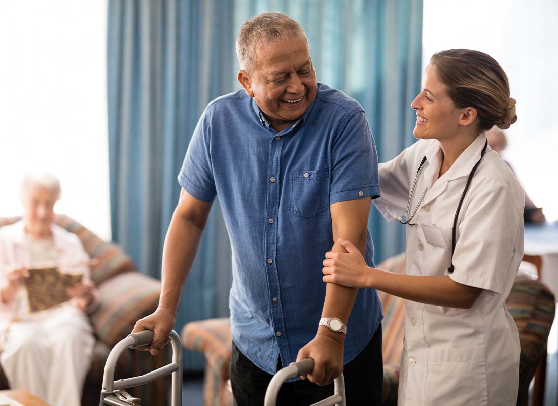 Senior Care Facility Insurance - Smiling Man With Walker Looking at a Female Caregiver Offering Physical Support as He tries to Make His Way to His Room at a Senior Care Facility